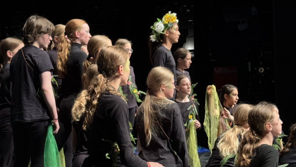 Szene aus einem aufgeführten Stück der jungen Truppe; ein Mädchen mit Blumenschmuck in den Haaren steht inmitten von vielen anderen schwarz gekleideten Kindern.