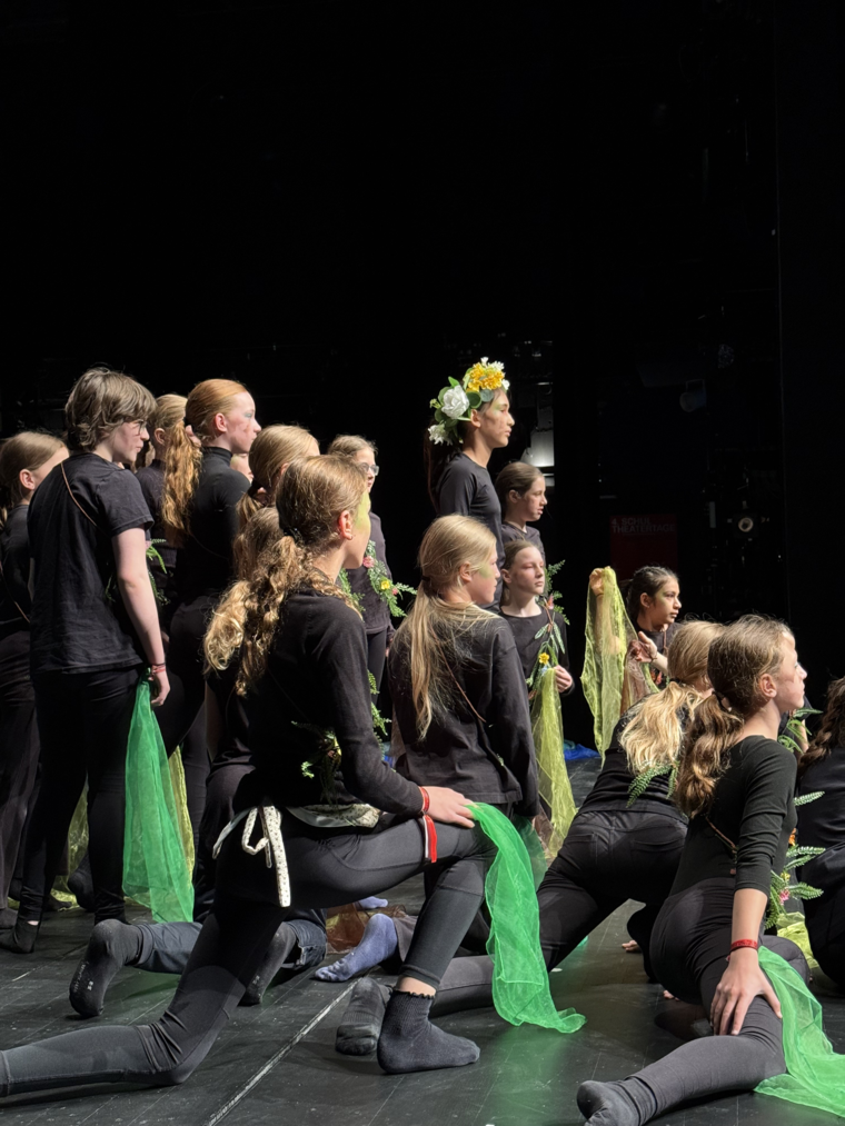 Szene aus einem aufgeführten Stück der jungen Truppe; ein Mädchen mit Blumenschmuck in den Haaren steht inmitten von vielen anderen schwarz gekleideten Kindern.