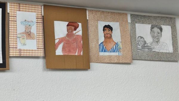 Fünf Zeichnungen hängen an einer Wand, die verschiedene Personen in unterschiedlichen Stilen und Farben darstellen.