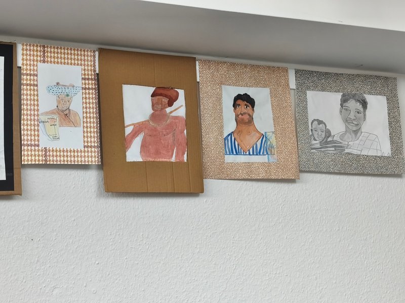 Fünf Zeichnungen hängen an einer Wand, die verschiedene Personen in unterschiedlichen Stilen und Farben darstellen.