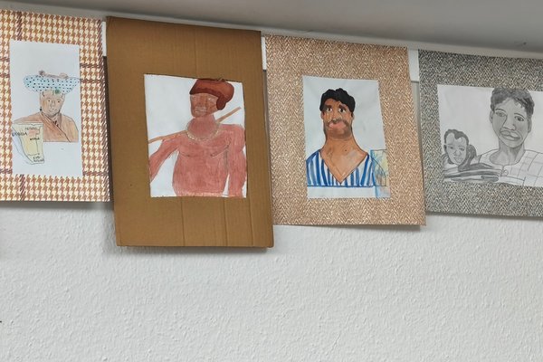 Fünf Zeichnungen hängen an einer Wand, die verschiedene Personen in unterschiedlichen Stilen und Farben darstellen.