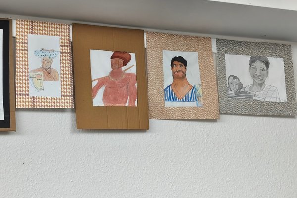 Fünf Zeichnungen hängen an einer Wand, die verschiedene Personen in unterschiedlichen Stilen und Farben darstellen.