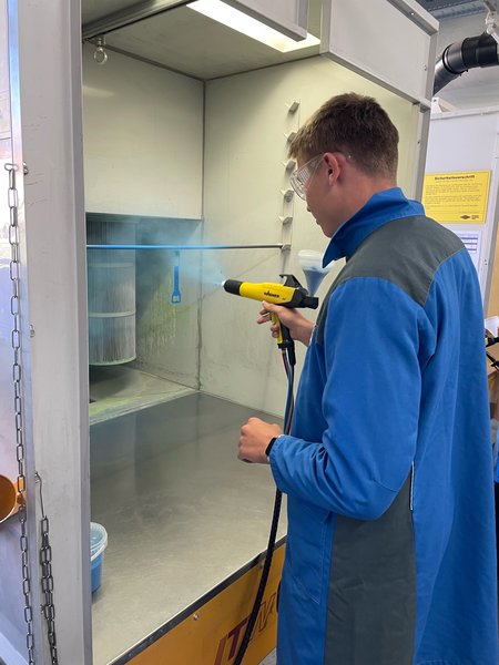 Eine Person in blauem Overall verwendet eine Sprühpistole in einem Reinigungsraum.